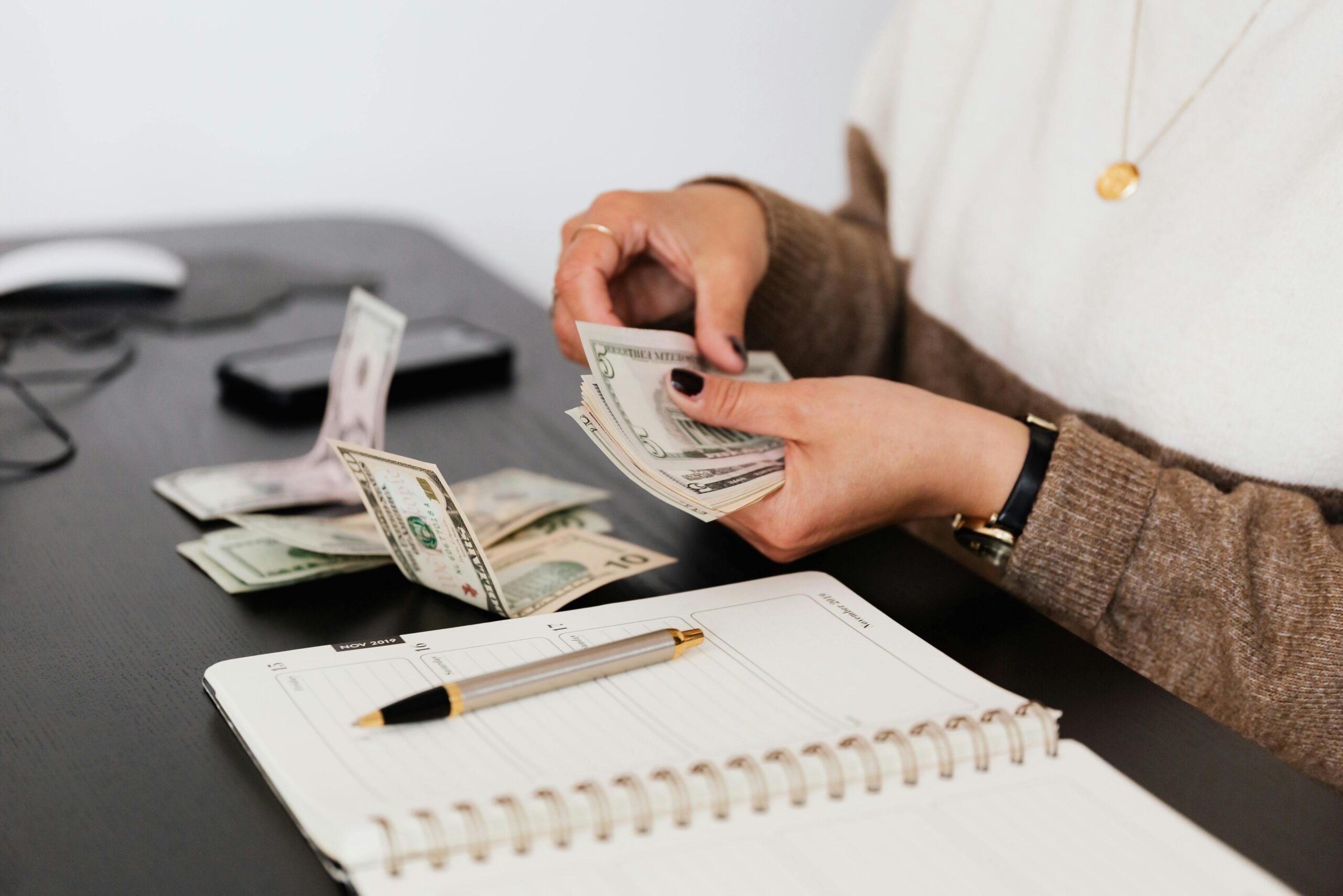 A person with painted nails flips through cash bills at their desk with a pen and paper nearby.