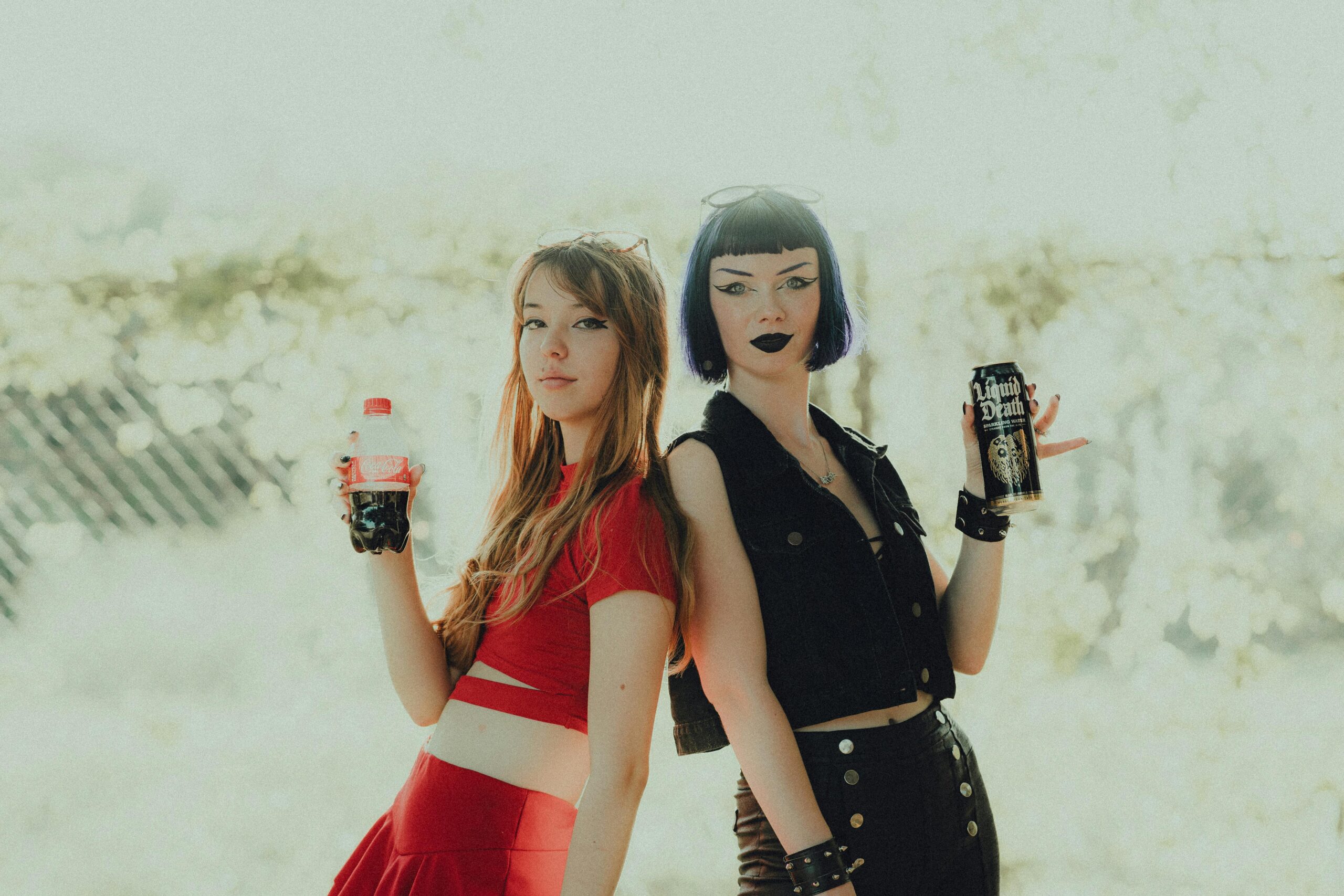 Two customers hold drinks, one dressed in red holing a coca-cola bottle, one in all black holding a Liquid Death can