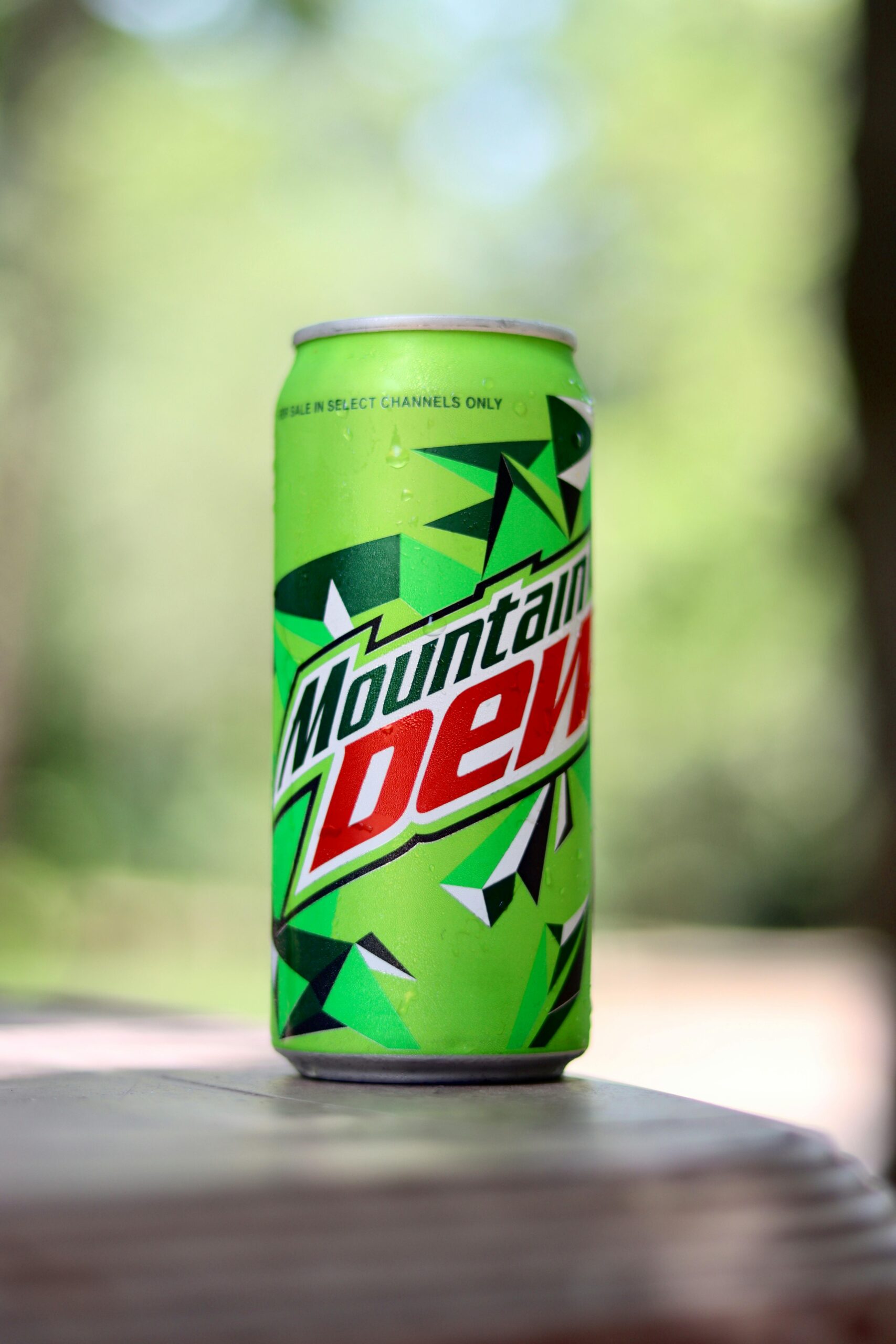 A can of bright green mountain dew sits on a table with some green trees out of focus in the background